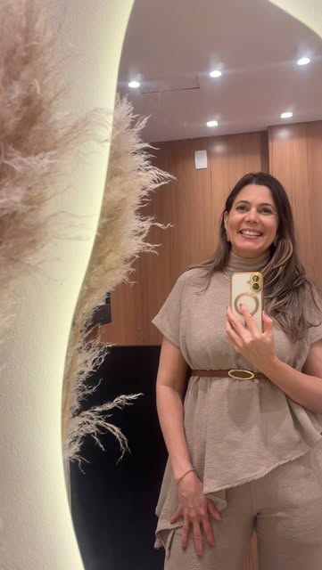 Simone Paixão sorrindo em frente ao espelho iluminado da clínica, de conjunto bege, com capim-dos-pampas ao lado.